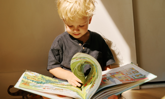 A young child sits on the floor in sunlight, reading a large, colorful picture book in their lap.